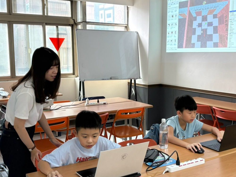 左營教室的 Roblox 營隊課堂，老師彎腰看學生的筆電畫面，隔壁同學專心操作，前方投影遊戲關卡。