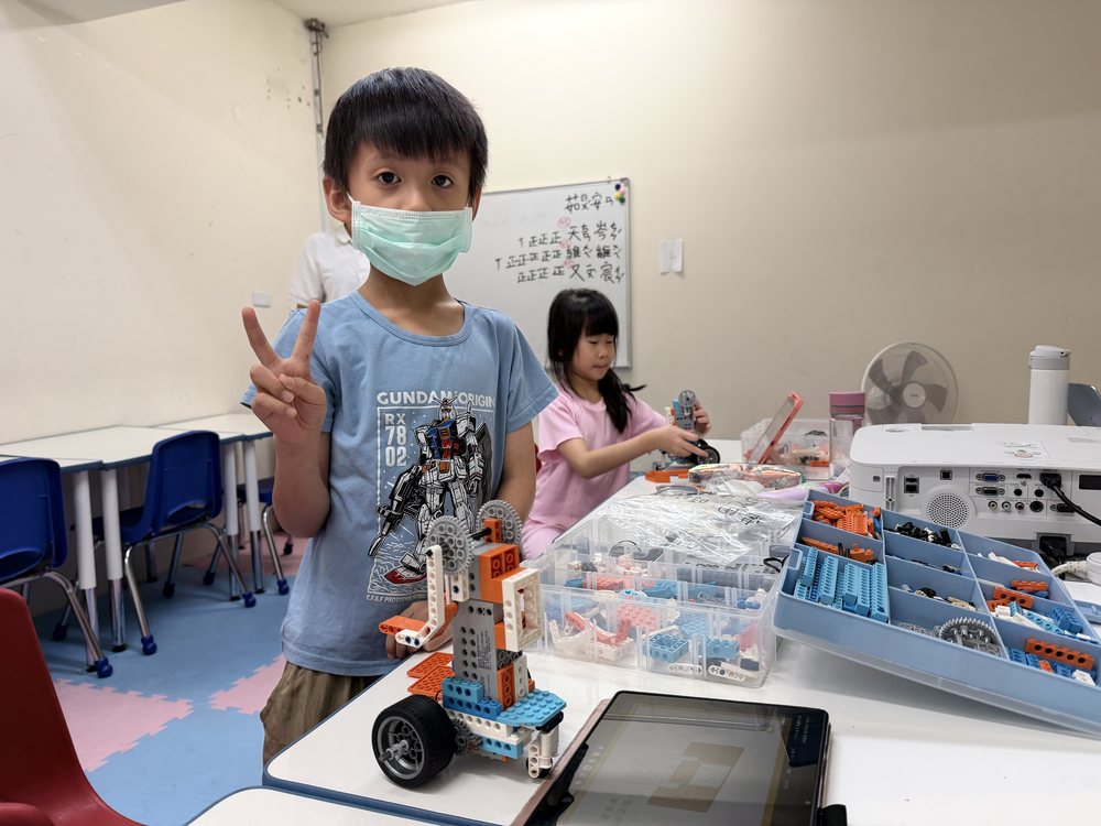 台中北屯教室 STEAM 創意機械積木課，學生完成自己組裝的機械車作品，桌上是零件盒與平板