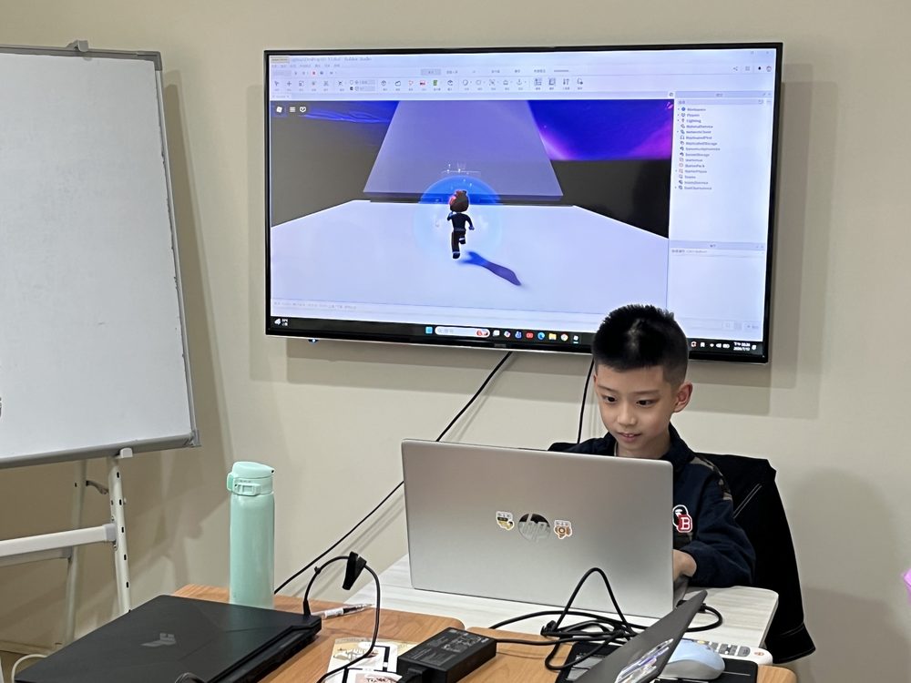 古亭教室的營隊課堂，學生坐在筆電前，把自己用 Roblox Studio 做的遊戲投到牆上大螢幕展示。