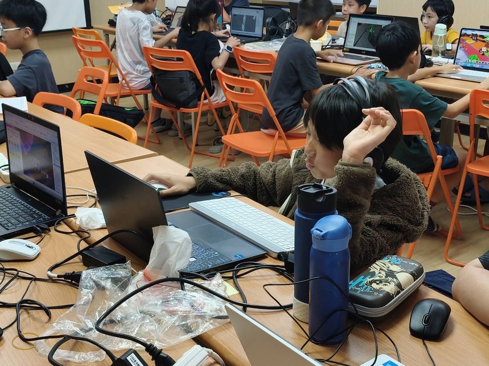 古亭教室的營隊課堂，學生們坐在長桌前各自使用筆電，螢幕上開著 Roblox Studio 製作遊戲場景。